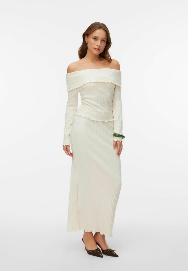 Balta, pleciem nokarāma, ribotā adīta kleita ar slāņotu kakla izgriezumu, garām piedurknēm un viļņotu apakšmalu, ko papildina tumši, smailu purngalu zābaki.