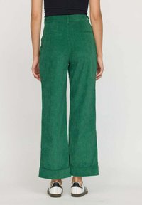 Pantalons en velours côtelé vert à jambe large avec une taille haute, dotés de poches arrière et de poignets retroussés. Associés à des chaussures blanches et noires.