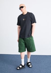 T-shirt noir en coton avec le logo Nike doré, shorts en lin verts, chaussettes blanches et sandales noires. Le modèle se tient devant un fond clair.