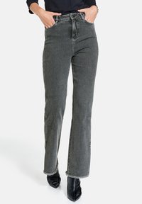 Pantalones vaqueros acampanados de mezclilla gris de cintura alta con dobladillo sin terminar, con bolsillos frontales y detalles de costura sutiles, combinados con botas negras de punta.