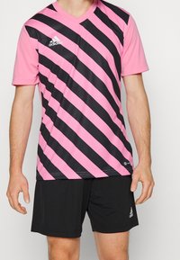 Maglia sportiva a maniche corte a righe rosa e nere con scollo a V. Indossata con pantaloncini neri, presenta il marchio Adidas.