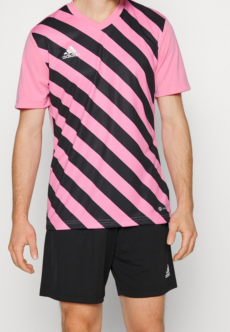 Maglia sportiva a maniche corte a righe rosa e nere con scollo a V. Indossata con pantaloncini neri, presenta il marchio Adidas.