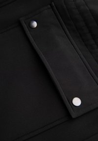 Tela negra con una textura suave, con una solapa rectangular y dos botones de presión metálicos. Costuras visibles a lo largo de los bordes.