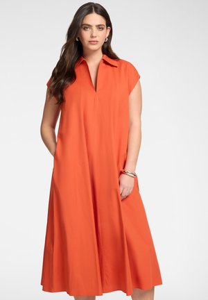 Kobieta z długimi ciemnymi włosami ubrana w pomarańczową sukienkę z krótkim rękawem, kołnierzem i kieszeniami oraz srebrne kolczyki koła i bransoletkę.