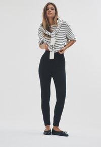 Fekete magasderekú leggings, fehér csíkos crop pulóverrel párosítva, amelynél sálra emlékeztető megkötő van a nyaknál, és fekete balettcipővel kiegészítve.