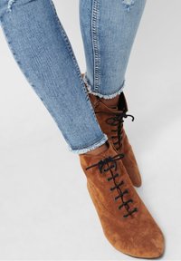 Smala ben i ljusblå jeans med fransar, iförda spetsiga bruna mocka snörkängor mot en ljusgrå bakgrund.