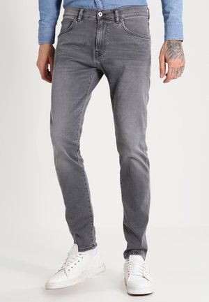 Mand iført slim fit grå jeans og hvide sneakers, stående mod en ensartet lys baggrund med synlig tatoveret hånd.