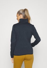 Merinovillainen tikkijakku, jossa on korkea kaulus, mustat olkapääpalkit ja muotoiltu malli, yhdistettynä sinapinkeltaisiin housuihin.