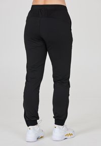 Sorte træningsbukser med tætsiddende pasform, elastisk talje og smalle ben, parret med hvide sneakers med gyldne detaljer.