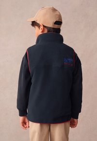 Tamsiai mėlynas flisinis švarkas su aukštu apykakle, raudonomis detalėmis ir išsiuvinėtu „SUPERDRY“ logotipu nugaroje, derinamas su smėlio spalvos kelnėmis ir smėlio spalvos kepure.