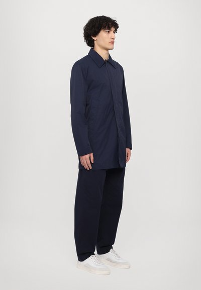 Manteau long bleu marine avec un col classique, coupe droite et fermeture à boutons, associé à un pantalon assorti et des baskets blanches.