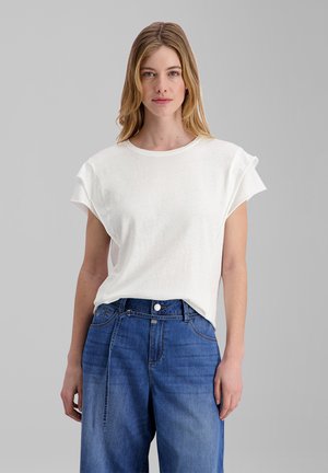 Young woman with shoulder-length blonde hair wears a white sleeveless top and high-waisted blue jeans against a plain gray background.