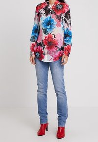 Blouse florale multicolore à manches longues, col à boutons et fond noir. Portée avec un jean bleu clair et des bottes à talons rouges.