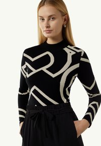 Schwarzer Strickpullover mit cremefarbenen abstrakten geometrischen Mustern, mit hohem Ausschnitt und langen Ärmeln. Kombiniert mit schwarzen Hosen mit Taillengürtel.