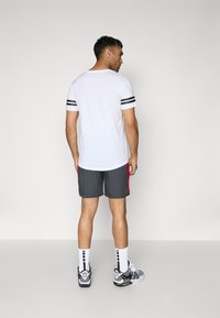 Hvid kortærmet skjorte med sorte striber på ærmerne, parret med grå shorts med røde accenter. Model iført sort og hvidt sneakers.
