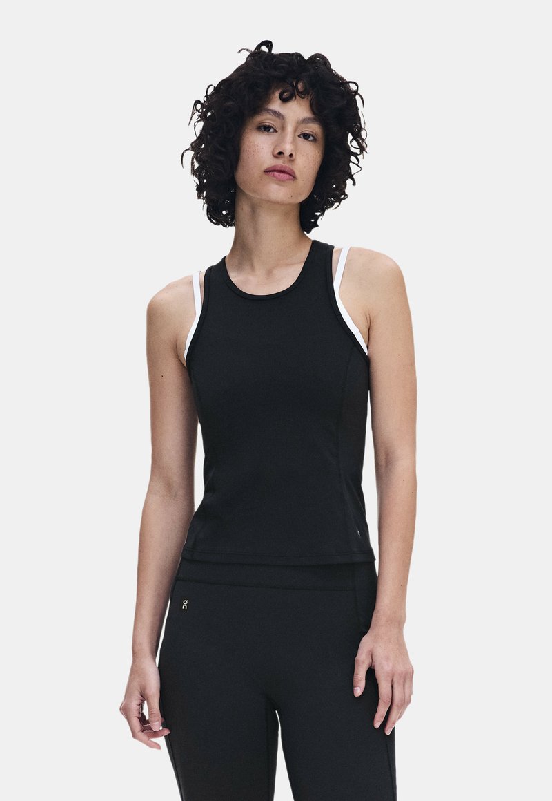 Femme aux cheveux bouclés foncés portant un haut de sport noir sans manches par-dessus un soutien-gorge de sport blanc et un pantalon de sport noir, debout devant un fond uni.