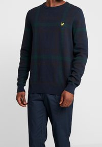 Maglione blu navy con un motivo a quadri verde e marrone, scollatura rotonda, maniche lunghe e un piccolo logo giallo sul petto.