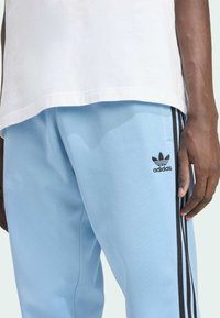 Valkoiset urheiluhousut, jotka on valmistettu pehmeästä kankaasta ja joissa on mustat sivutarrat sekä Adidas-logo mustana, esitetty valkoisen paidan kanssa.
