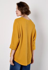 Haut jaune à texture gaufrée avec un grand col et des manches trois-quarts, présentant une coupe décontractée et des fentes sur les côtés pour plus de confort.
