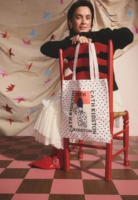 Oboustranná látková taška s puntíkovým vzorem a grafickým potiskem. Jasně červené detaily a pevné ramenní popruhy. Na pozadí dřevěná židle.