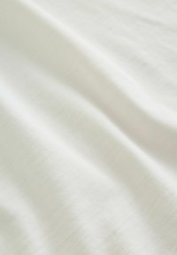 Tissu blanc doux avec une texture tissée subtile, présenté en plis délicats créant des variations de lumière et d’ombre.