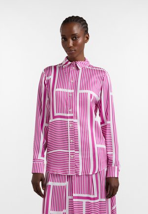 Frau trägt ein langärmeliges rosa-weiß gestreiftes Hemd mit Knöpfen und einem passenden Rock, steht vor einem schlichten weißen Hintergrund.