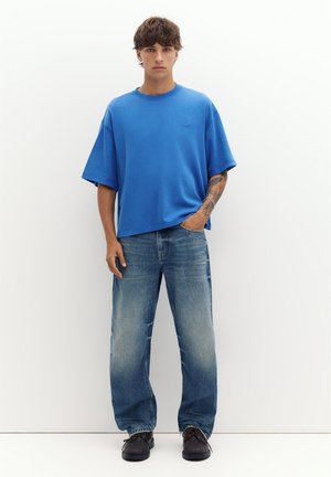 Młody mężczyzna stoi, ubrany w oversize niebieski T-shirt, luźne sprane dżinsy, czarne buty, z tatuażem i ręką w kieszeni, neutralne tło.