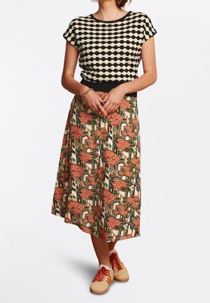 Frau trägt ein schwarz-weiß gemustertes Oberteil, einen floralen geometrischen Midirock und beige Sneakers mit orangen Schnürsenkeln, steht mit verschränkten Händen.