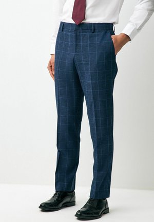 Pantalons de costume à carreaux bleu foncé en tissu texturé, avec une coupe ajustée et des poches latérales, associés à des chaussures de ville noires et une chemise blanche.