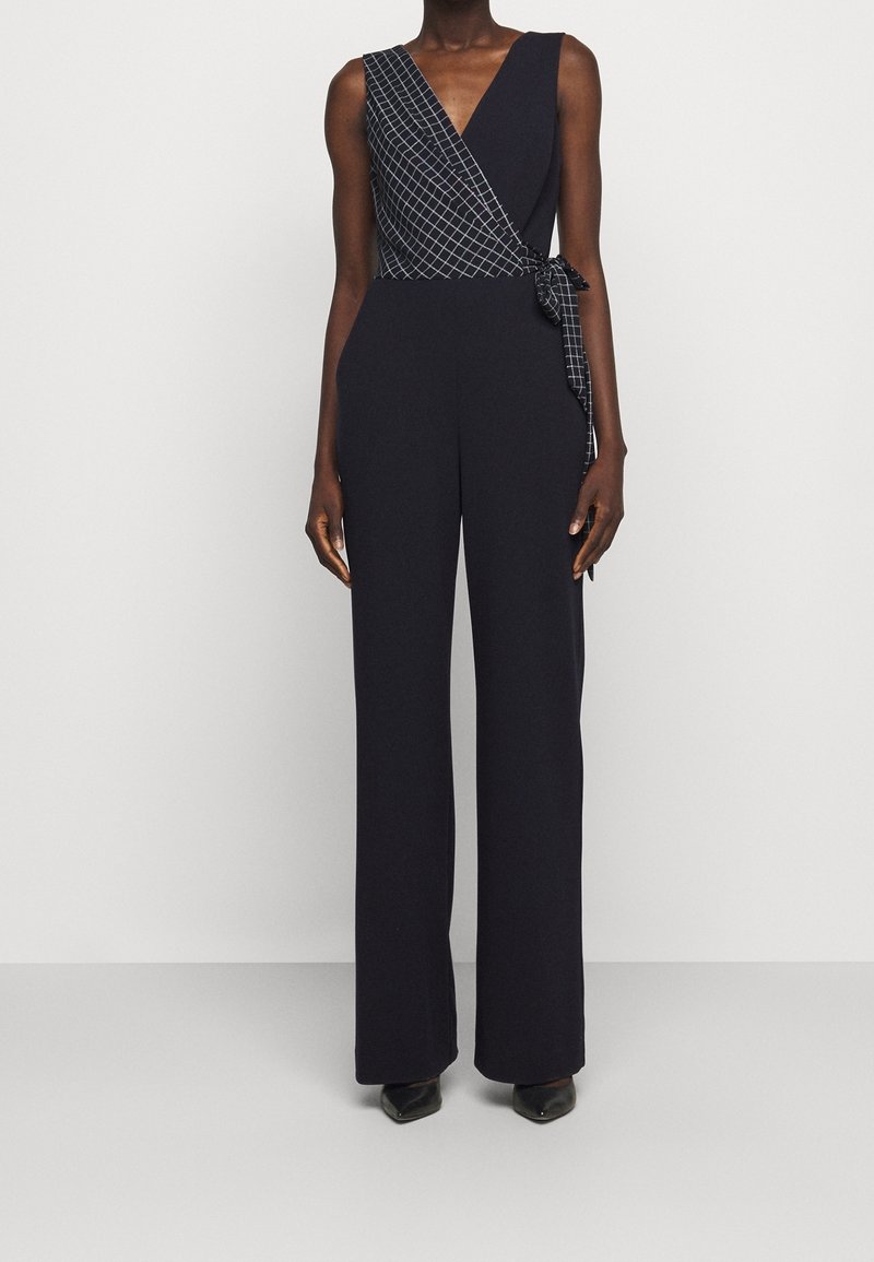 Laivastonsininen haalarimuotoilu, jossa on kääntöosa, sisältäen musta-valkoisen geometrisen kuviopanelin ja vyötä. Sujuva kangas, leveät lahkeet.