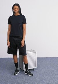 Svart bomull t-skjorte, denimshorts og sorte sneakers med hvite detaljer; står ved en sølvfarget lagringskasse med tekstur på et blått teppe.