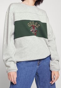 Šedá mikina s zeleným vodorovným pruhem, na kterém je vyšitý text "Guess" a barevný erb. Materiál z bavlněné směsi, volný střih.