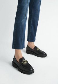 Mocassini in pelle nera con suola spessa, caratterizzati da un dettagli di fibbia dorata. Abbinati a jeans in denim blu a taglio corto con orli grezzi.
