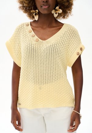 Kobieta ubrana w bladożółty, dziergany krótki top z drewnianymi guzikami na dekolcie i rękawach, w połączeniu z jasnymi spodniami.