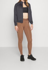 Mörkgrå atletisk zip-jacka, svart sportbh, bruna leggings och svarta sneakers. Tight passform med sömlösa design detaljer.