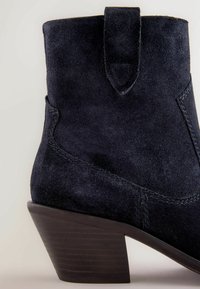 Stivaletti alla caviglia in suede blu scuro con punta affusolata, tacco in legno impilato e dettaglio con asola laterale. Le cuciture visibili aggiungono texture e design.