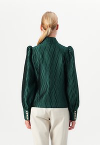 Woman with blonde ponytail wearing a dark green textured long-sleeve blouse with puffed shoulders and beige pants, shown from the back.
