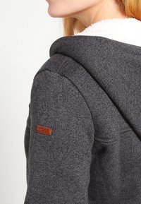 Grå huvtröja med ett mjukt, krämfärgat foder, med en läderlogotyp på vänster axel. Texturerad tyg och figurnära design.