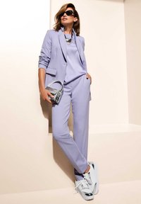 Lavendelfarbenes Anzug mit tailliertem Blazer und maßgeschneiderter Hose, kombiniert mit einem hellen Oberteil und silbernen Sneakers. Akzentuiert mit einer metallischen Handtasche und Sonnenbrille.