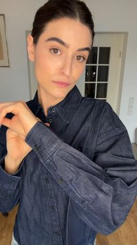 Žena s tamnom kosom koja nosi tamnu traper košulju, gleda u kameru dok prilagođava lijevi rukav u zatvorenom prostoru.