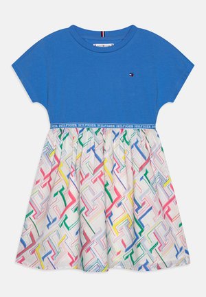 Niebieska bawełniana bluzka z krótkimi rękawami, skontrastowana kolorową, wzorzystą spódnicą z geometrycznymi kształtami w odcieniach różu, żółci, niebieskiego i zielonego.