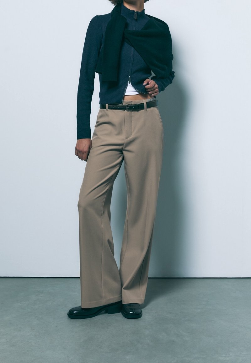 Personne portant un pantalon beige à jambes larges, ceinture noire, haut zippé bleu marine avec un pull noir posé sur les épaules, et chaussures noires, debout contre un mur blanc.