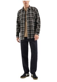 Karierter Button-up-Hemd in Schwarz, Braun und Weiß, über einem beigen Grafik-T-Shirt getragen; kombiniert mit dunklen Jeans und schwarzen Sneakers.