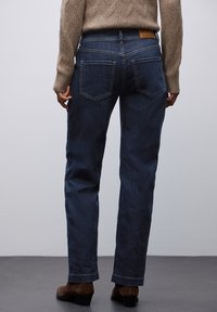 Jeans de mezclilla azul oscuro con corte recto, que cuentan con dos bolsillos traseros y una etiqueta de cuero en la cintura. Combinados con botas marrones de tobillo.