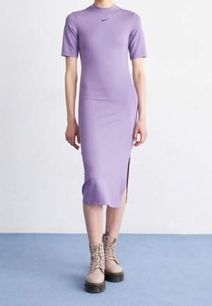 Personne portant une robe midi lavande ajustée avec manches courtes, fente latérale, logo Nike noir et bottines beiges à lacets sur un sol bleu.