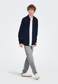 Mørkeblå knappeskjorte med to frontlommer, kombinert med hvit T-skjorte, grå jeans og hvite joggesko. Modell som går på ensfarget bakgrunn.