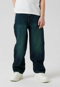 Tamno plave traperice s opuštenim krojem, denimski materijal, petlje za remen, pet džepova i blago izbjeljivanje; uparen s bijelim tenisicama.