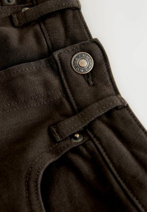 Tamsiai rudos spalvos džinso audinio artimas vaizdas, matyti metalinis mygtukas su įrašu „New Denim“, siūlių dygsniai ir audinio kilpa reguliavimui.