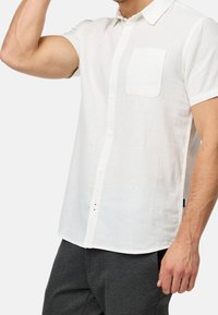 Mężczyzna w białej koszuli z krótkim rękawem i kieszonką na piersi, w ciemnoszarych spodniach, unosi lewą rękę w okolicach głowy.