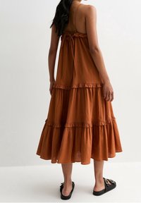 Femme portant une robe midi sans manches couleur rouille à volants avec des fines bretelles et des sandales à enfiler noires, se tenant devant un fond blanc uni.